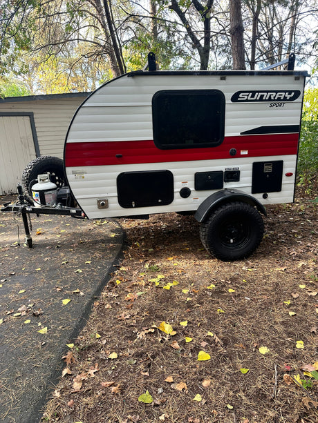 White & red Sunray Sport teardrop **rv rental**, with rugged tires, parked on a leafy drive. Perfect for adventure!
