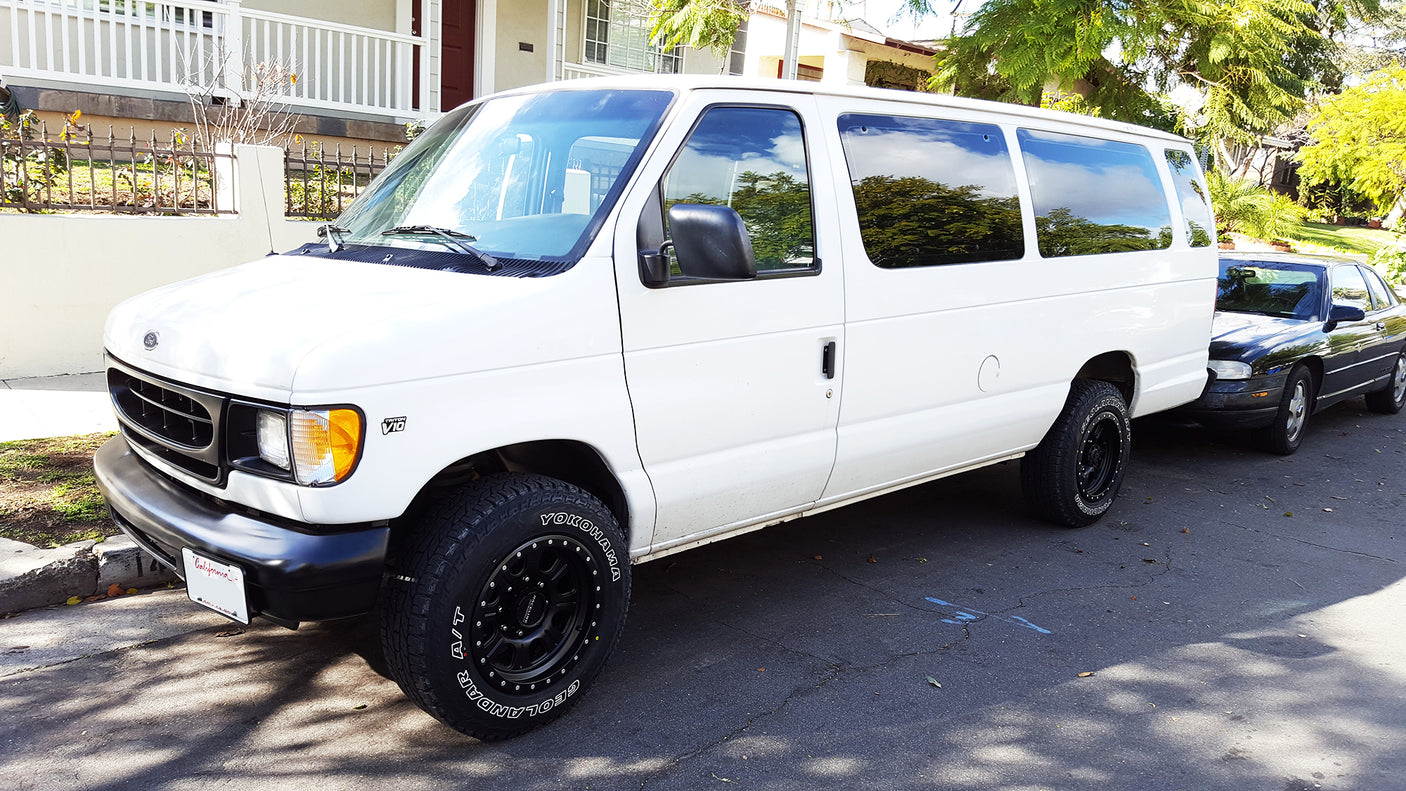 A white Ford V10 van with rugged tires, parked on a street in front of a black car. Could be a great rv rental.