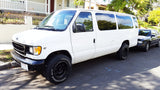 A white Ford V10 van with rugged tires, parked on a street in front of a black car. Could be a great rv rental.