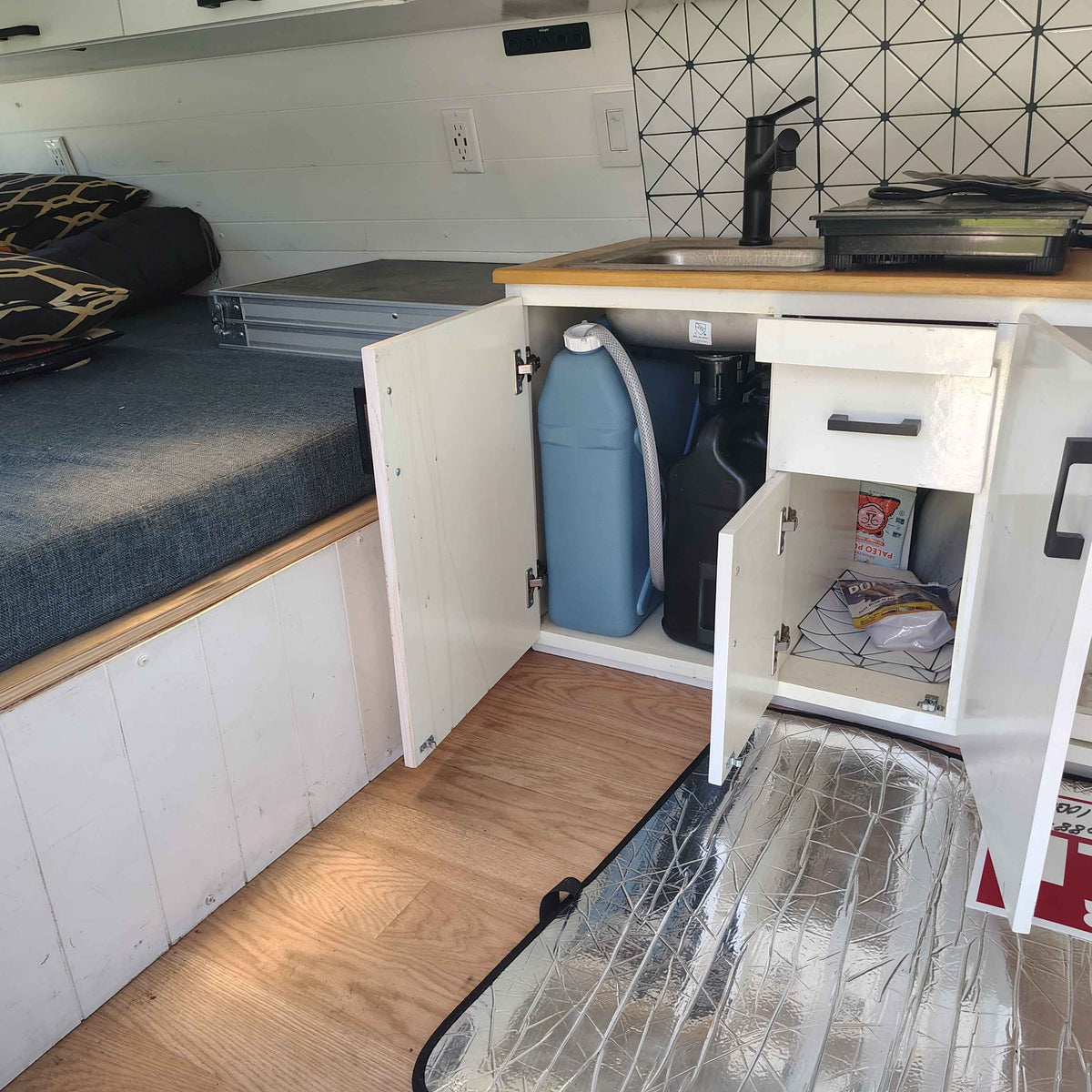 Inside an RV rental, featuring a bed, kitchen with sink/cooktop, and water storage containers in open cabinets. — photo 9