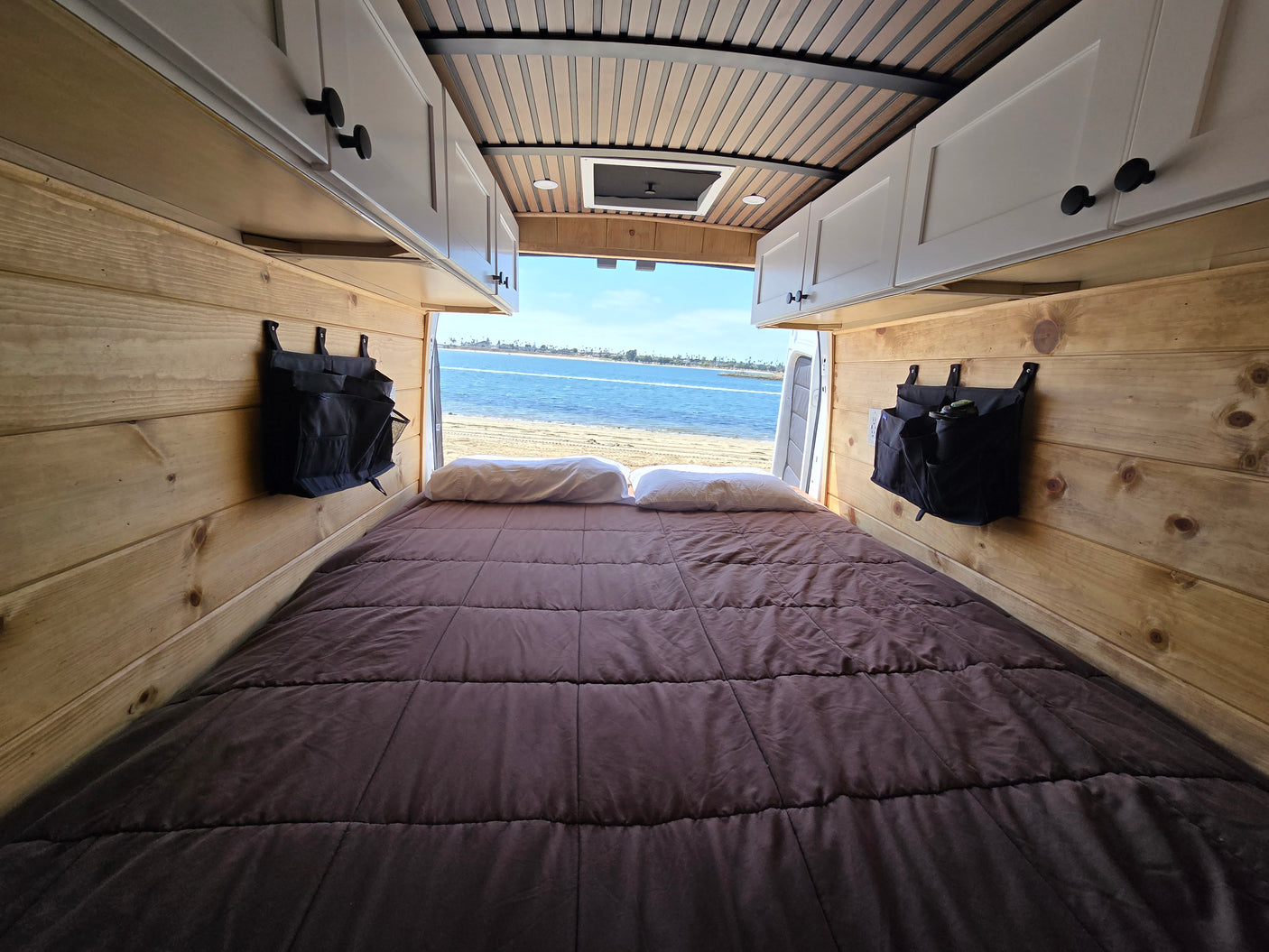 Cozy RV rental camper van interior: a bed with wood walls opens to a beautiful beach and ocean view.