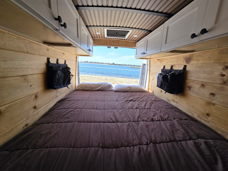  parked on a sandy shore overlooking a calm bay on a sunny day.|RV rental: Rear view of a camper van's wood-paneled interior — photo 3