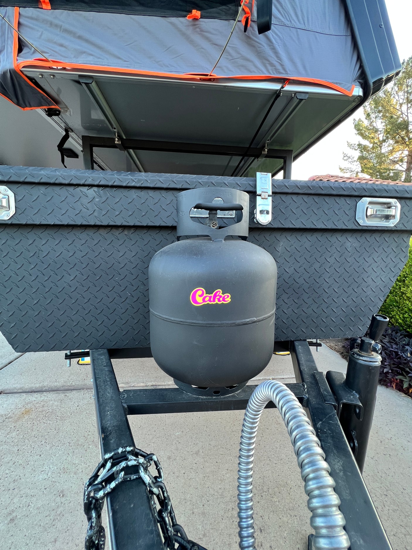 This RV rental trailer features a front diamond plate storage box, a black propane tank, and a grey rooftop tent.