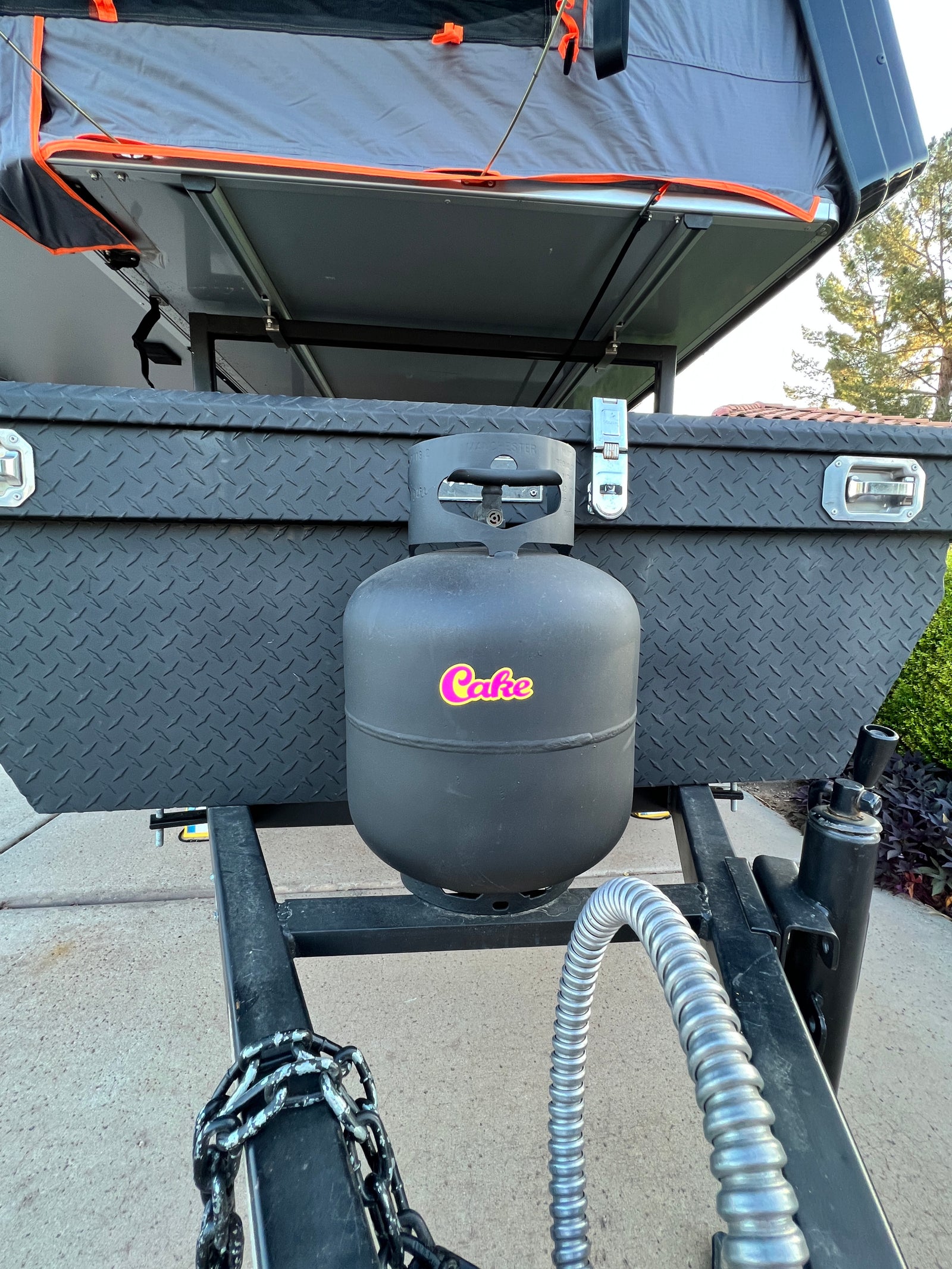 This RV rental trailer features a front diamond plate storage box, a black propane tank, and a grey rooftop tent.