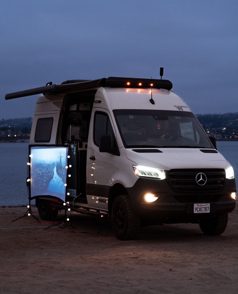  surrounded by tall trees.|This RV rental is parked on a sandy beach with its rear doors open — photo 8