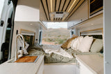 Inside a spacious RV rental: cozy bed, sink, and cabinets, with a desert landscape visible through the rear doors. Ideal for van life.