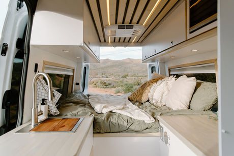 Inside a spacious RV rental: cozy bed, sink, and cabinets, with a desert landscape visible through the rear doors. Ideal for van life.