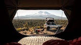 From inside a tent, a white SUV with a rooftop tent, two people, and a dog are camping by a mountain. Perfect for your next **RV rental** **adventure**!
