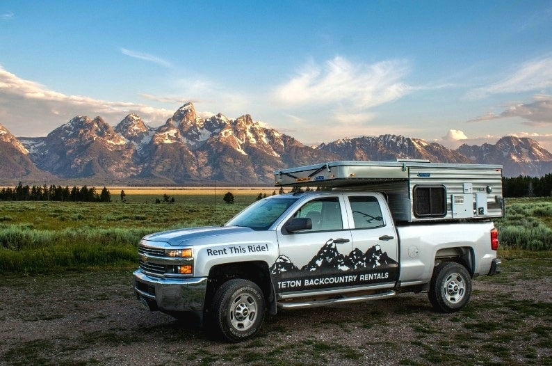 Explore the Tetons with this RV rental truck camper. Silver pickup parked with mountains & green fields in the background. — photo 5