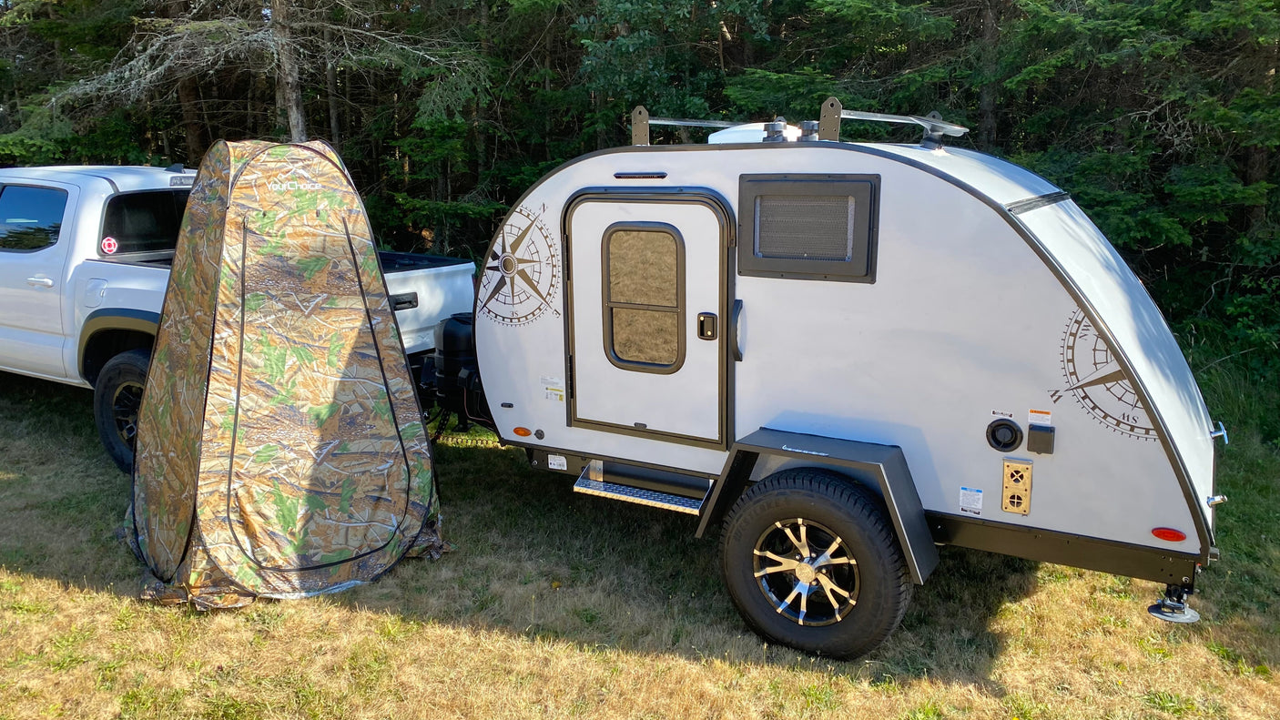 RV rental ready: A light grey teardrop camper and camouflage privacy tent beside a white truck in a sunny, grassy area.