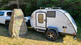 RV rental ready: A light grey teardrop camper and camouflage privacy tent beside a white truck in a sunny, grassy area.