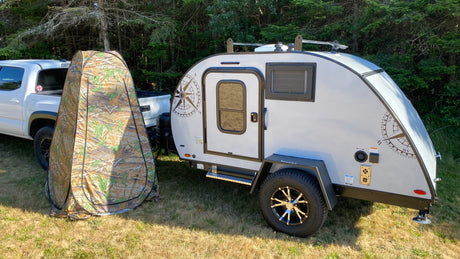 RV rental ready: A light grey teardrop camper and camouflage privacy tent beside a white truck in a sunny, grassy area.