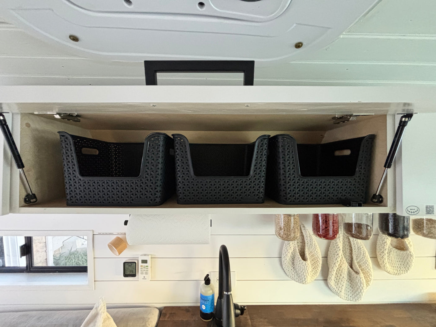 Open overhead cabinet with storage baskets in an RV rental kitchen, featuring a sink, paper towels, and hanging spice jars.