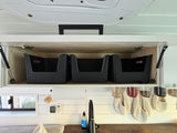 Open overhead cabinet with storage baskets in an RV rental kitchen, featuring a sink, paper towels, and hanging spice jars.