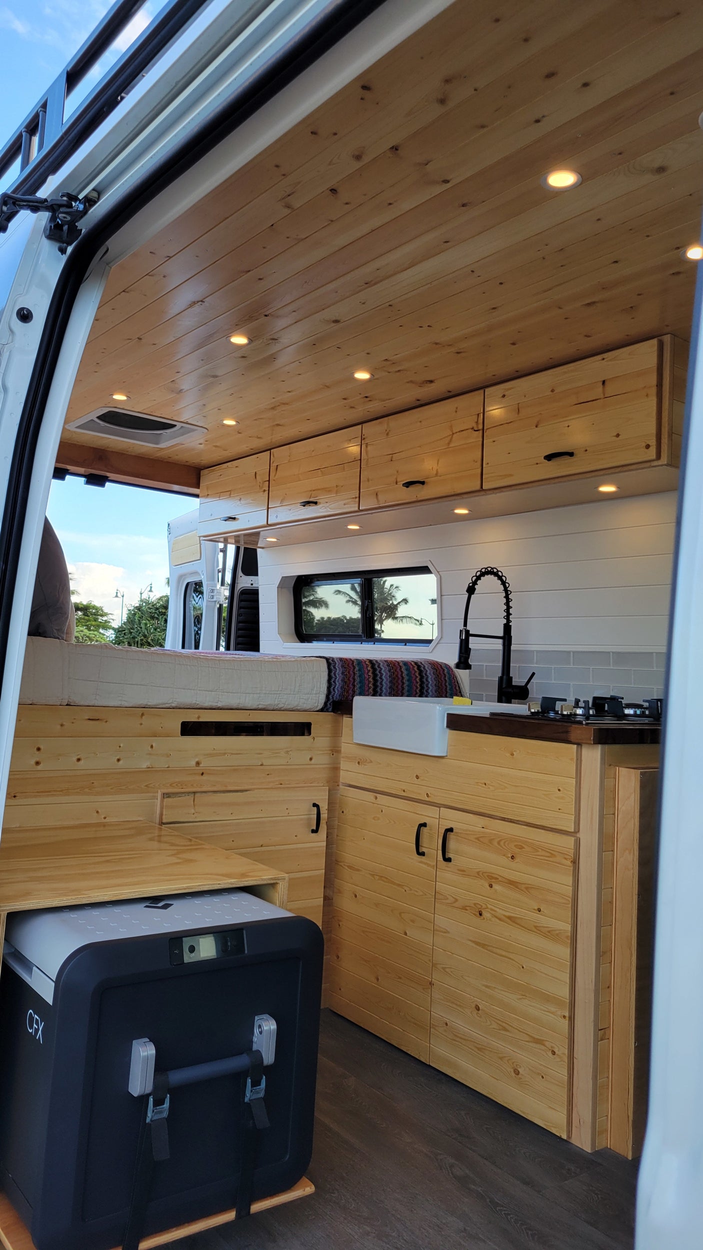 RV rental interior featuring a bright wood-paneled ceiling, comfortable bed, compact kitchenette, and a Dometic CFX cooler.