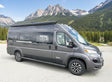 Grey RV rental camper van parked against a scenic mountain range and pine forest under a blue sky