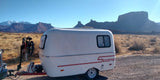 This rv rental shows a compact white Scamp trailer with a bicycle attached, set against a wide desert landscape and snow-capped mountains.