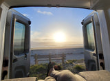 Enjoying the beach sunset from an RV rental. Open doors offer a relaxing view of the ocean, sand, and bench from inside the camper.
