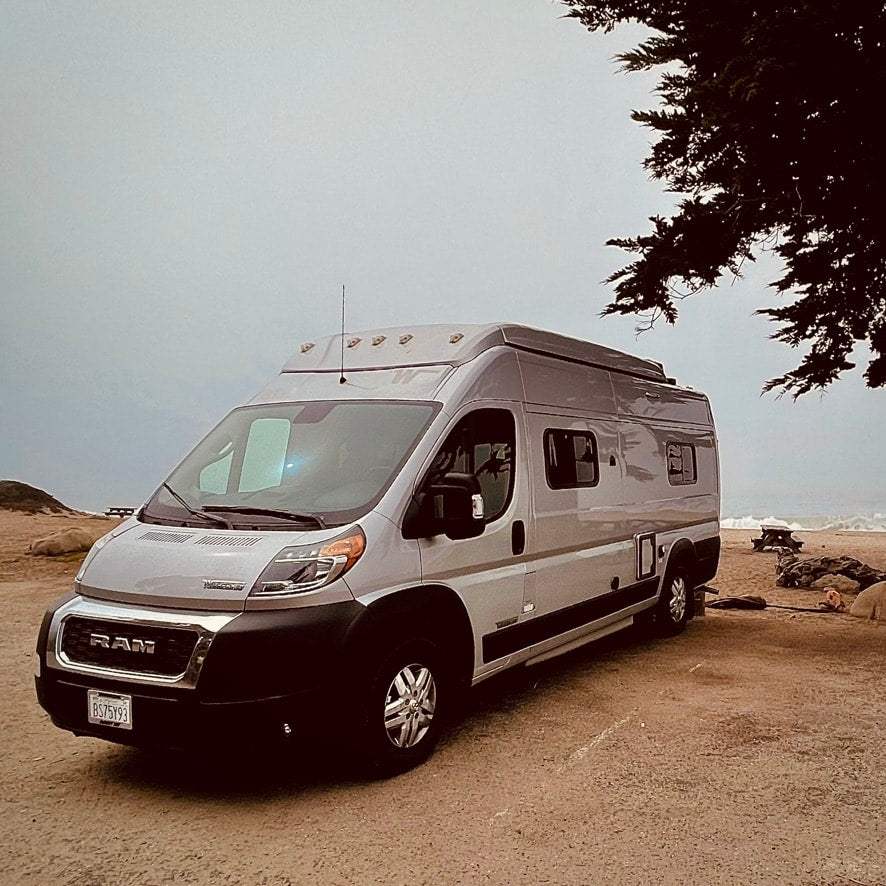 An RV rental camper van parked on sand by a misty ocean, under a tree. Perfect for beach adventures.