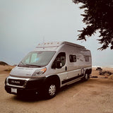 An RV rental camper van parked on sand by a misty ocean, under a tree. Perfect for beach adventures.