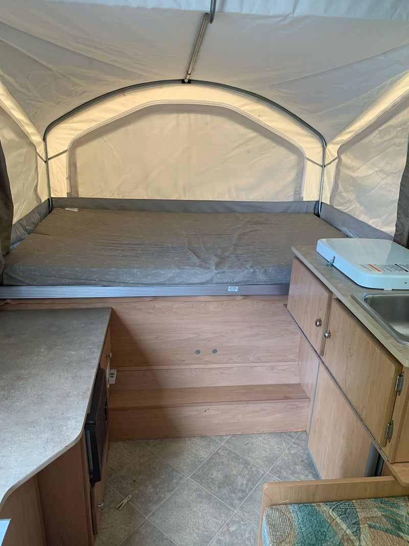 RV rental pop-up camper interior: extended bed, small kitchen with sink/stove, and wooden steps. Compact & functional. — photo 7