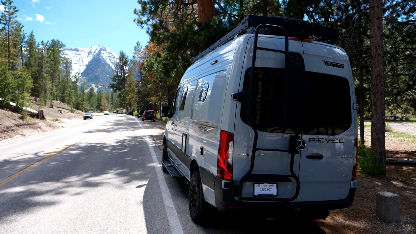 A Winnebago Revel rv rental parked on a mountain road with snow-capped peaks and pine trees. Great for a scenic road trip!