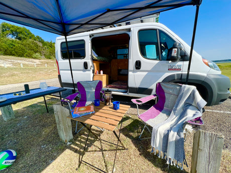 RV rental parked with a shaded outdoor setup, featuring chairs, coffee, books, and a blanket for relaxation.