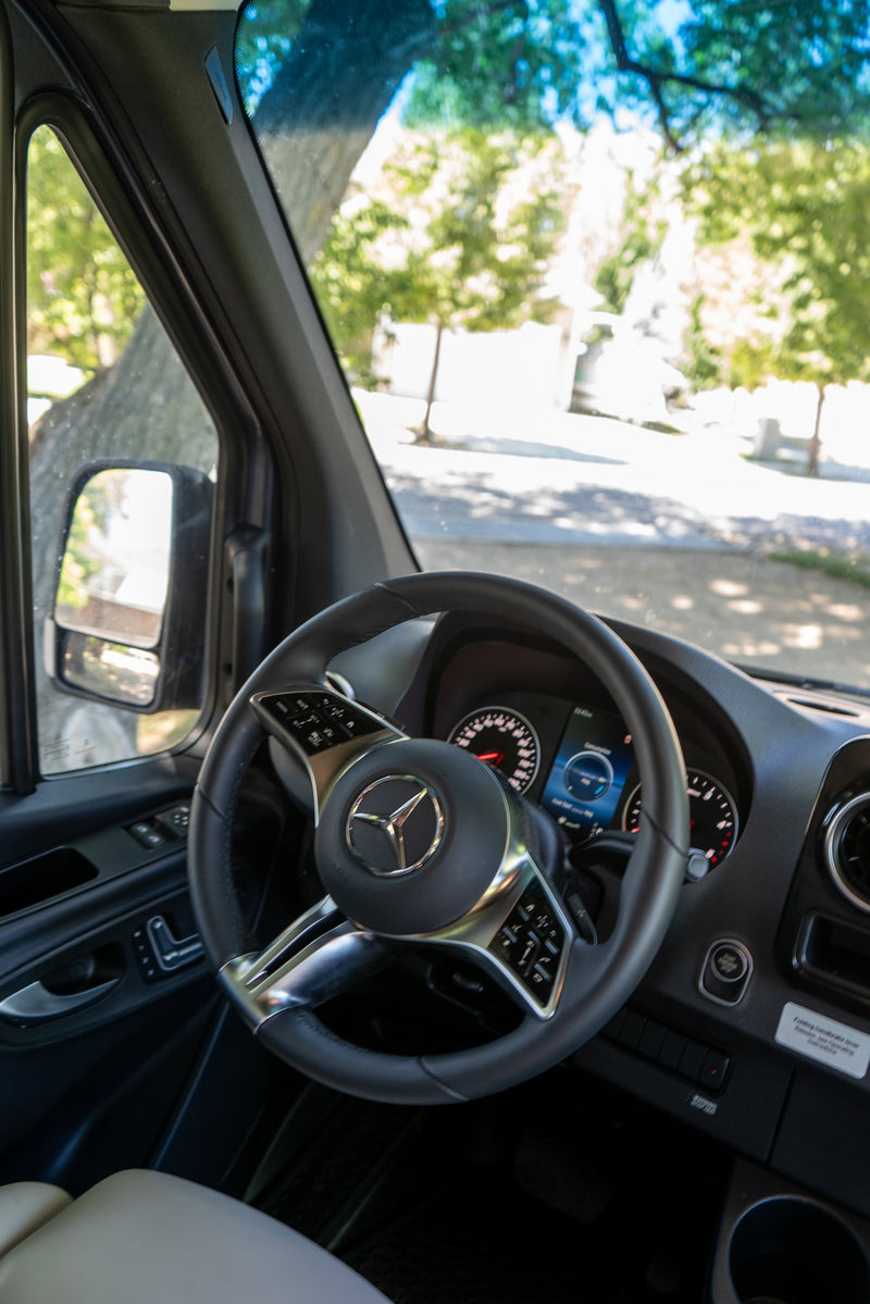 Inside an RV rental: a Mercedes-Benz steering wheel, digital dashboard, and a view of a sunny, treelined road. — photo 10