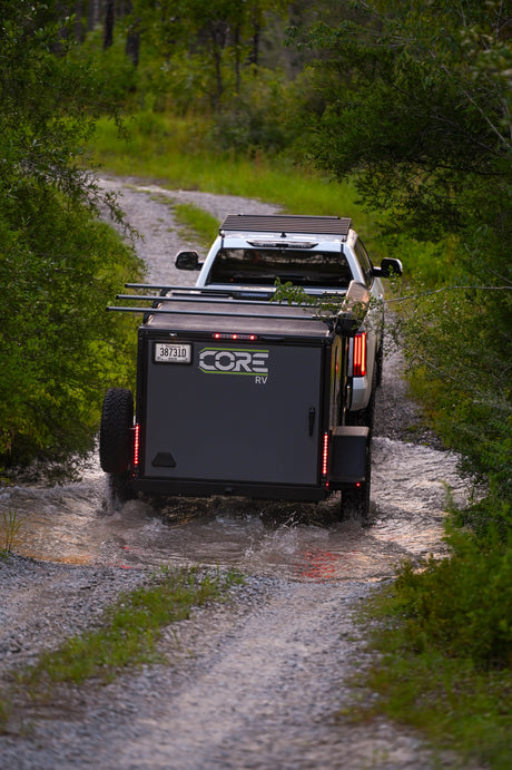 A CORE RV rental trailer, towed by a pickup, navigates a water crossing on a rugged gravel path, ideal for off-road adventure.