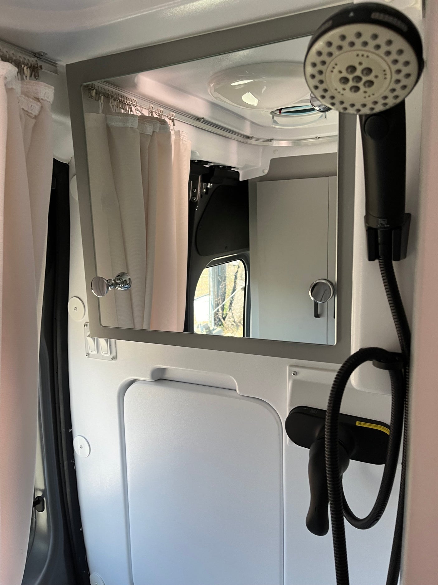 RV rental bathroom: sleek, compact space with hanging shower, mirror, and curtain. Reflection shows outside trees.