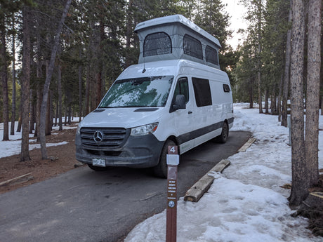 This white pop-top camper van, an RV rental, is parked at snowy campsite #4 in a forest.