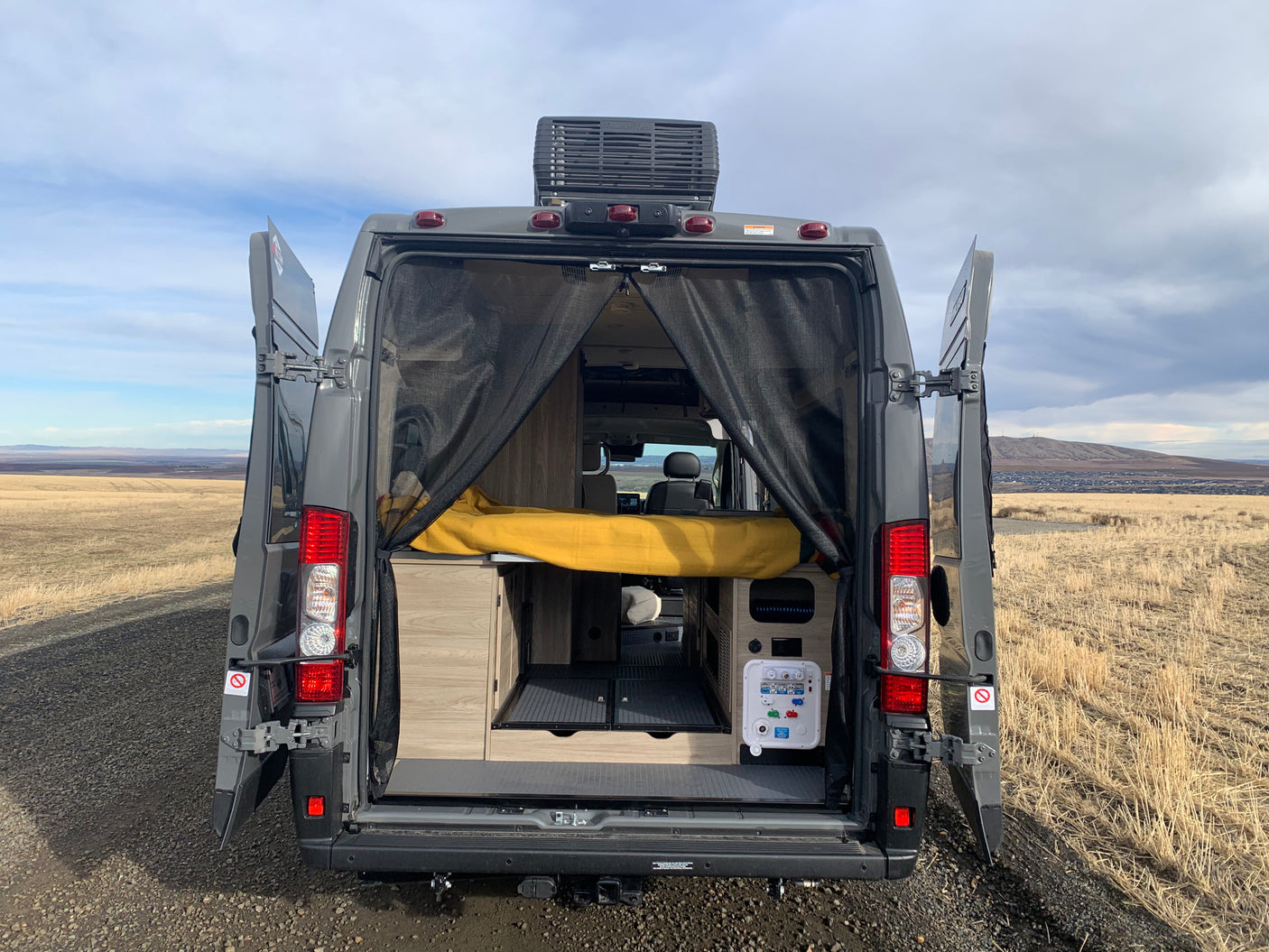 Open rear doors of a grey camper van RV, revealing a cozy interior with bed & utility panel. Ideal for an RV rental adventure.