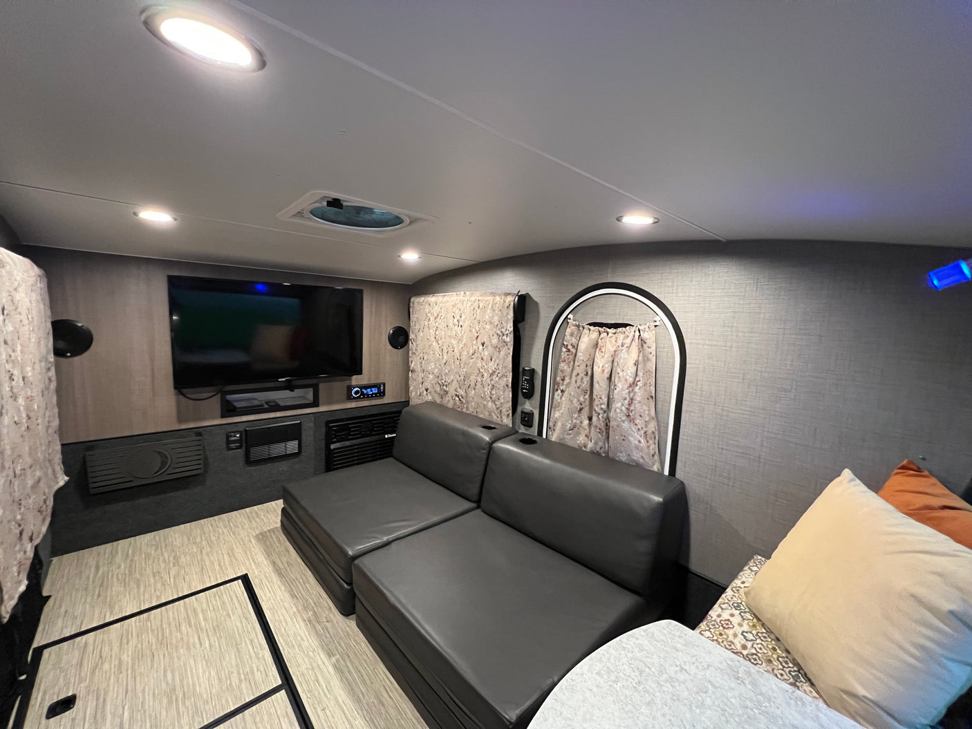 Spacious RV rental interior: view of the living area with a dark gray sectional sofa, wall-mounted TV, and bright lighting.
