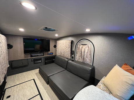 Spacious RV rental interior: view of the living area with a dark gray sectional sofa, wall-mounted TV, and bright lighting.
