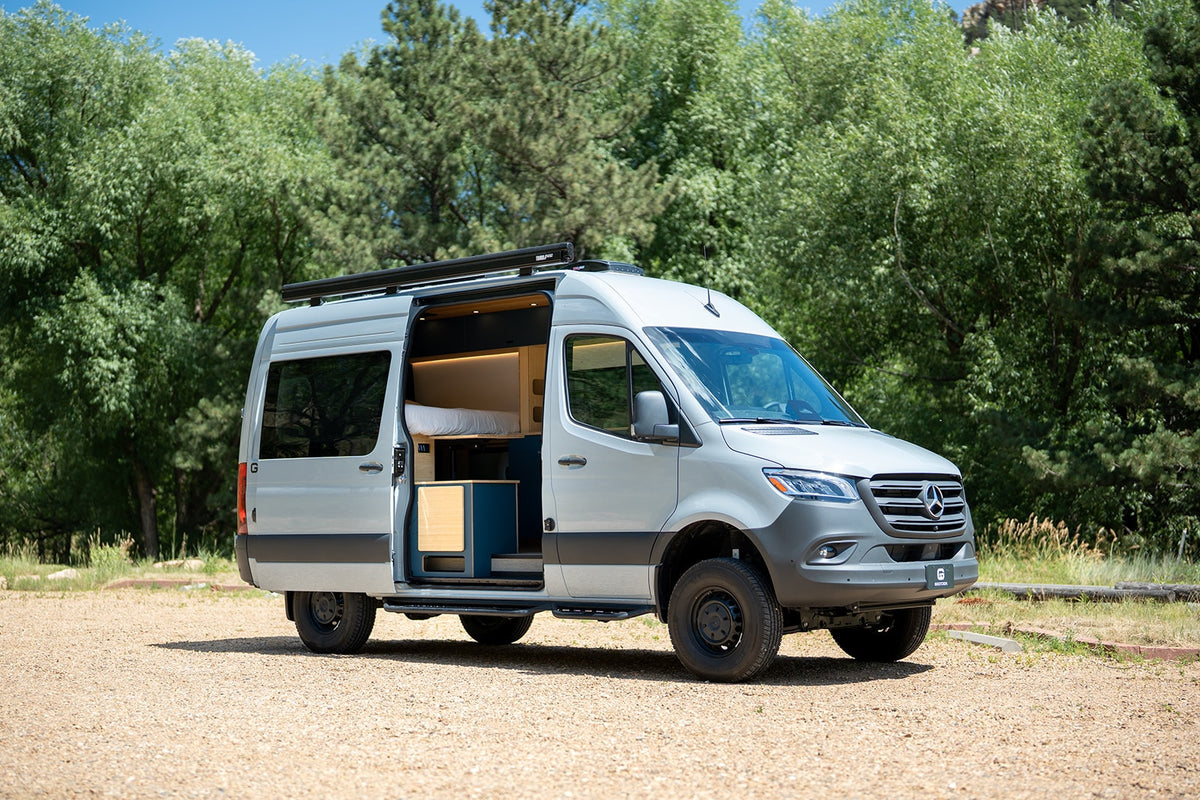 Spacious light gray camper van, perfect for rv rental trips. Open door reveals cozy interior, parked on gravel by trees.