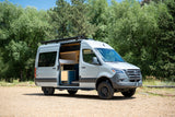 Spacious light gray camper van, perfect for rv rental trips. Open door reveals cozy interior, parked on gravel by trees.