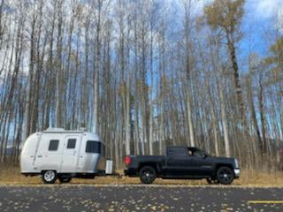 Black truck towing a white travel trailer on a road, flanked by tall, bare trees.