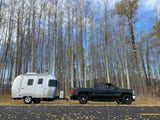 Black truck towing a white travel trailer on a road, flanked by tall, bare trees.