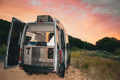 Stunning sunset over an RV rental camper van with its rear doors open, revealing a cozy interior ready for adventure.