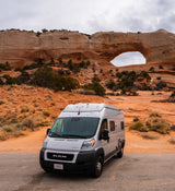 A silver RV rental camper van is parked in a vast red rock desert, with a majestic natural arch in the background on an overcast day.