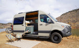 Experience nature with this rv rental camper van parked lakeside. Its open door reveals a cozy interior, with chairs & bike outside.