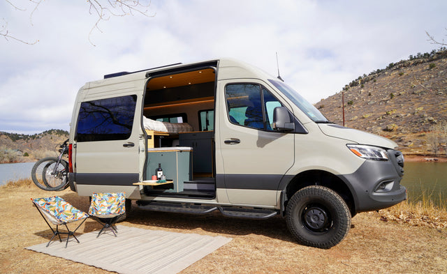Experience nature with this rv rental camper van parked lakeside. Its open door reveals a cozy interior, with chairs & bike outside.