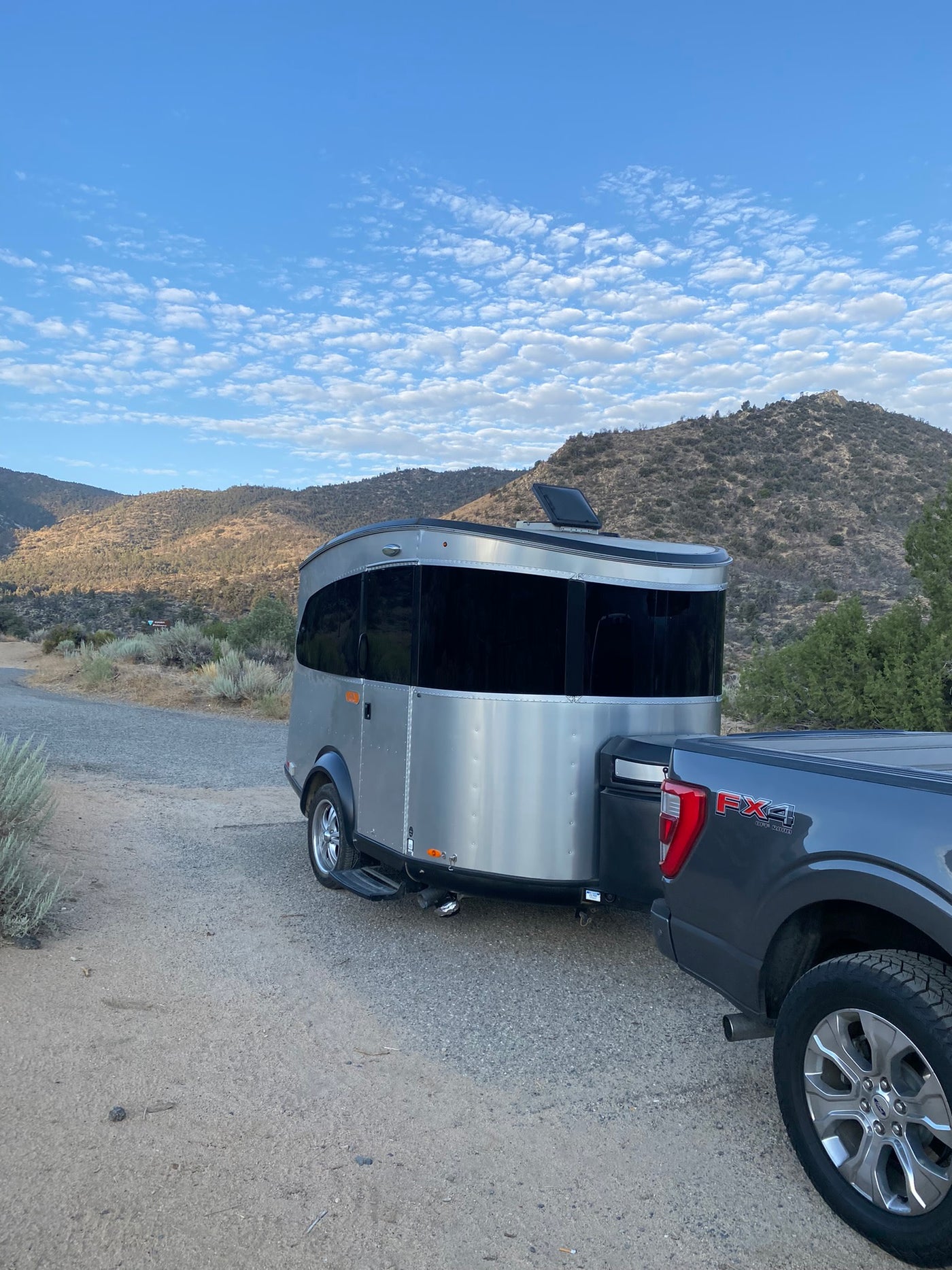 A silver compact RV trailer is hitched to a gray pickup truck on a dirt road, set against mountains. Perfect for an RV rental adventure. — photo 14