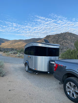 A silver compact RV trailer is hitched to a gray pickup truck on a dirt road, set against mountains. Perfect for an RV rental adventure.
