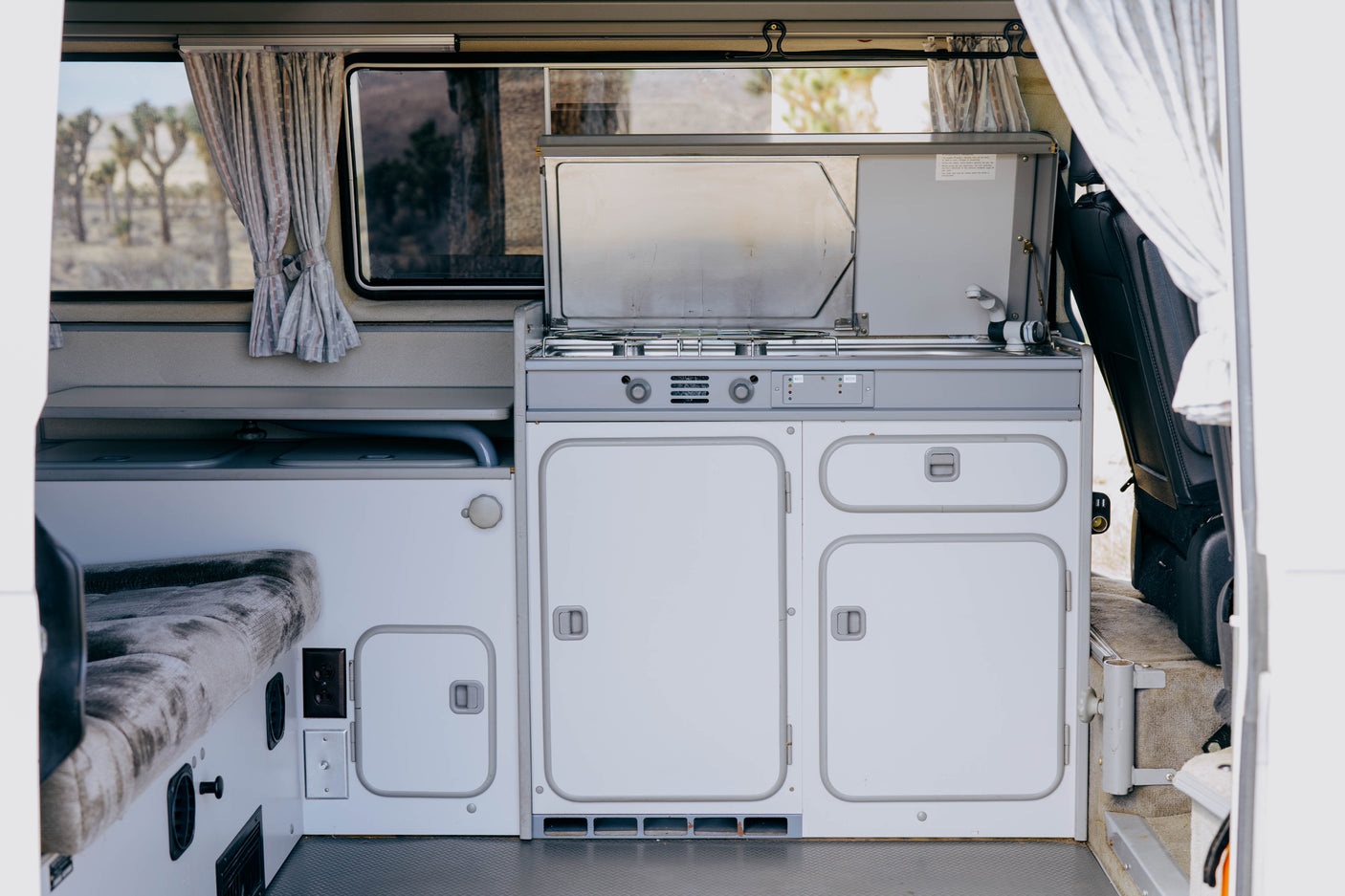 Explore this RV rental's camper van interior. Features a compact kitchen (stove, sink), bench, and desert views.