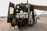 Two people work from a custom rv rental in a desert. Open back doors reveal bed, storage. An awning provides shade.