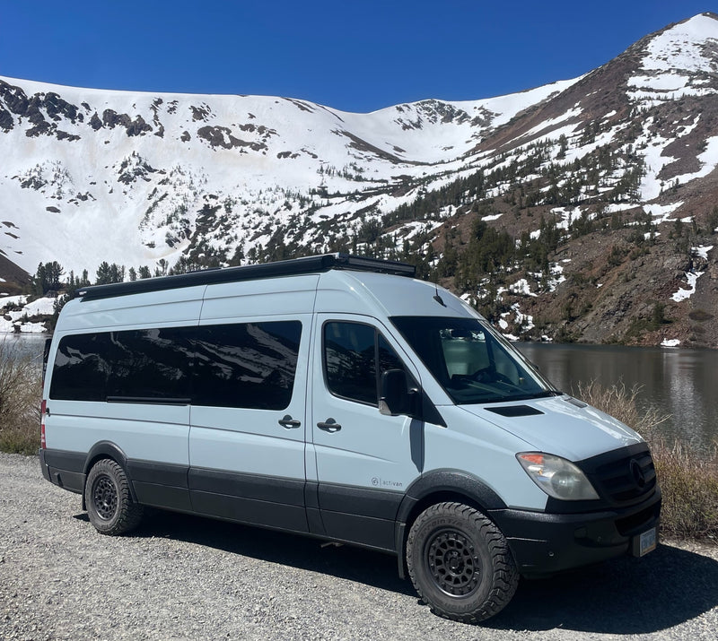 A light grey Sprinter rv rental camper van sits on a gravel road by a lake. Snow-covered mountains rise in the background.|Modern RV rental interior: a comfy bed — photo 1