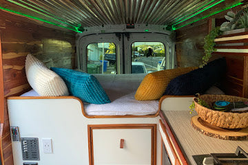 Step inside this RV rental! Cozy camper van interior with a bed, pillows, wood paneling, and green overhead lighting.