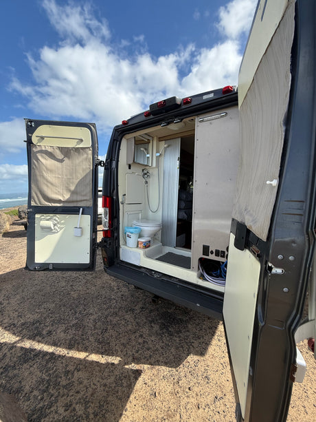 RV rental: Open back reveals a compact shower and toilet, perfect for camping. Parked by the ocean under blue skies.