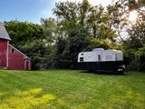 This vintage 'Urban Glamper' RV rental sits in a sunny green yard beside a rustic red barn, surrounded by lush trees.